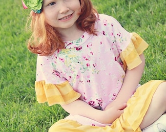 May include: A young girl with red hair wearing a pink and yellow floral dress with a green and blue flower headband. She is sitting on a grassy lawn.
