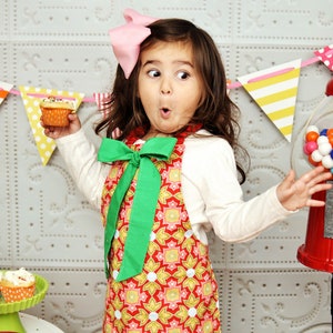 May include: A young girl wearing a red, yellow, and green patterned apron with a green bow, holding a cupcake with sprinkles. She is looking at a gumball machine with a surprised expression.
