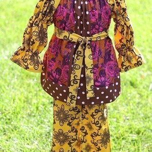 May include: A two-piece children's outfit featuring a tunic top and pants. The top has a scoop neck, long sleeves, and a tie belt. The outfit is a mix of brown, yellow, and pink floral patterns with white polka dots. The pants have a yellow floral design.