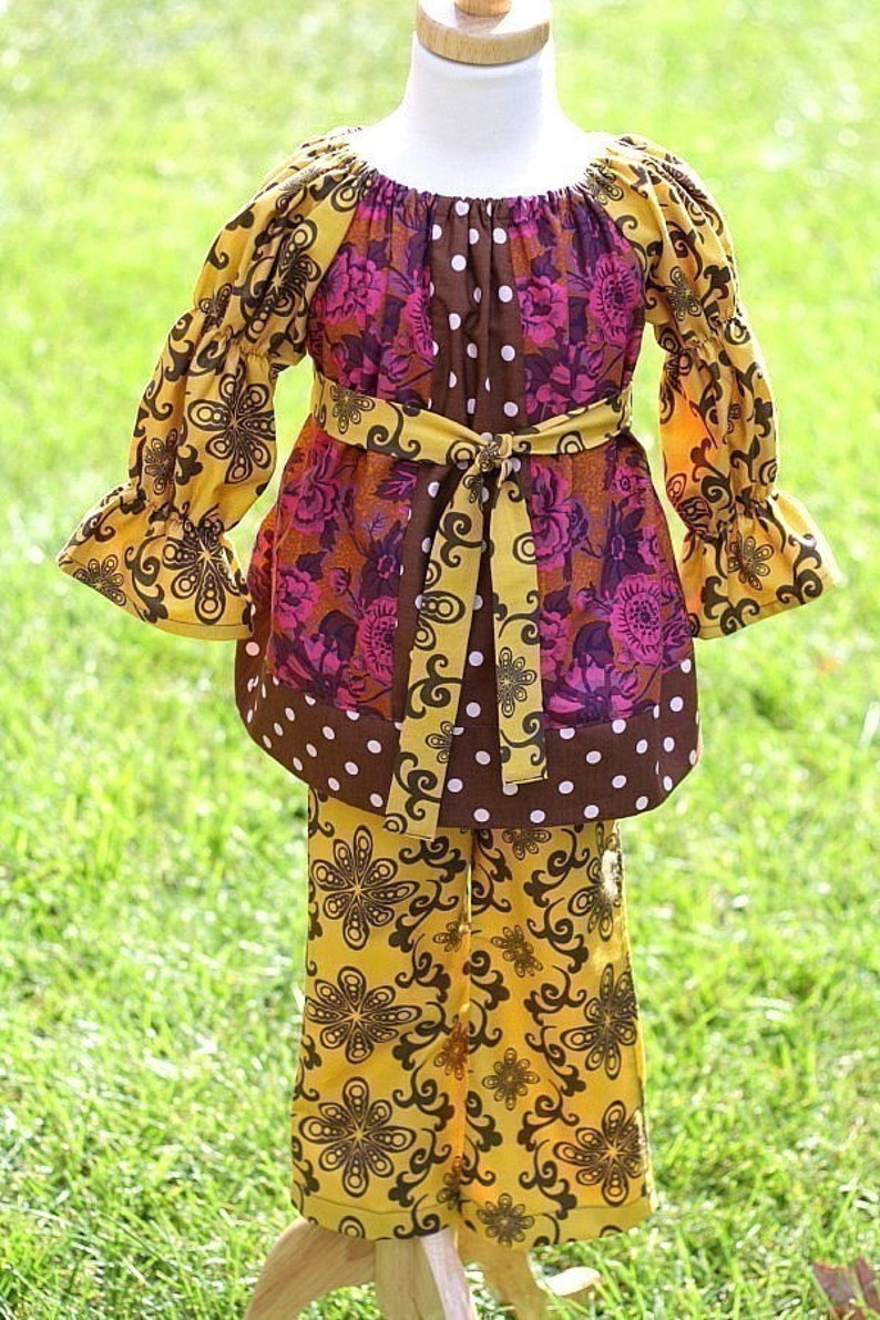 May include: A two-piece children's outfit featuring a tunic top and pants. The top has a scoop neck, long sleeves, and a tie belt. The outfit is a mix of brown, yellow, and pink floral patterns with white polka dots. The pants have a yellow floral design.