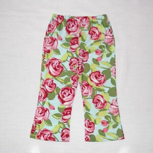 May include: A pair of pants with a floral pattern. The pants feature a light blue background with a repeating pattern of pink roses and green leaves. The pants have an elastic waistband and a straight leg.