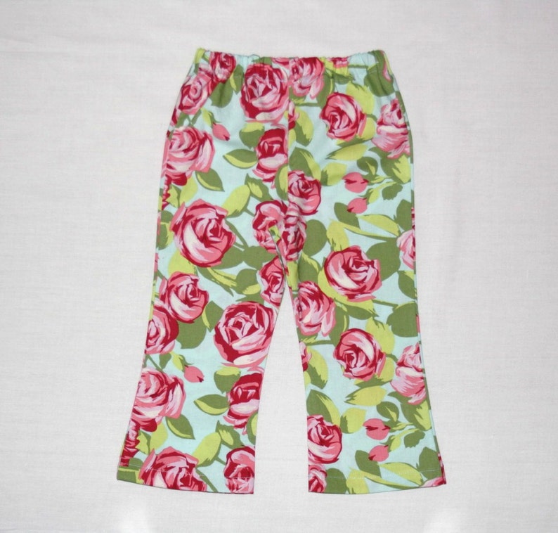 May include: A pair of pants with a floral pattern. The pants feature a light blue background with a repeating pattern of pink roses and green leaves. The pants have an elastic waistband and a straight leg.