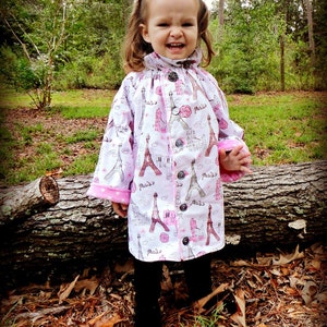 May include: A young girl wearing a white and pink jacket with a Paris theme. The jacket has a button closure and a pink trim. The girl is standing in front of a fallen log in a wooded area.