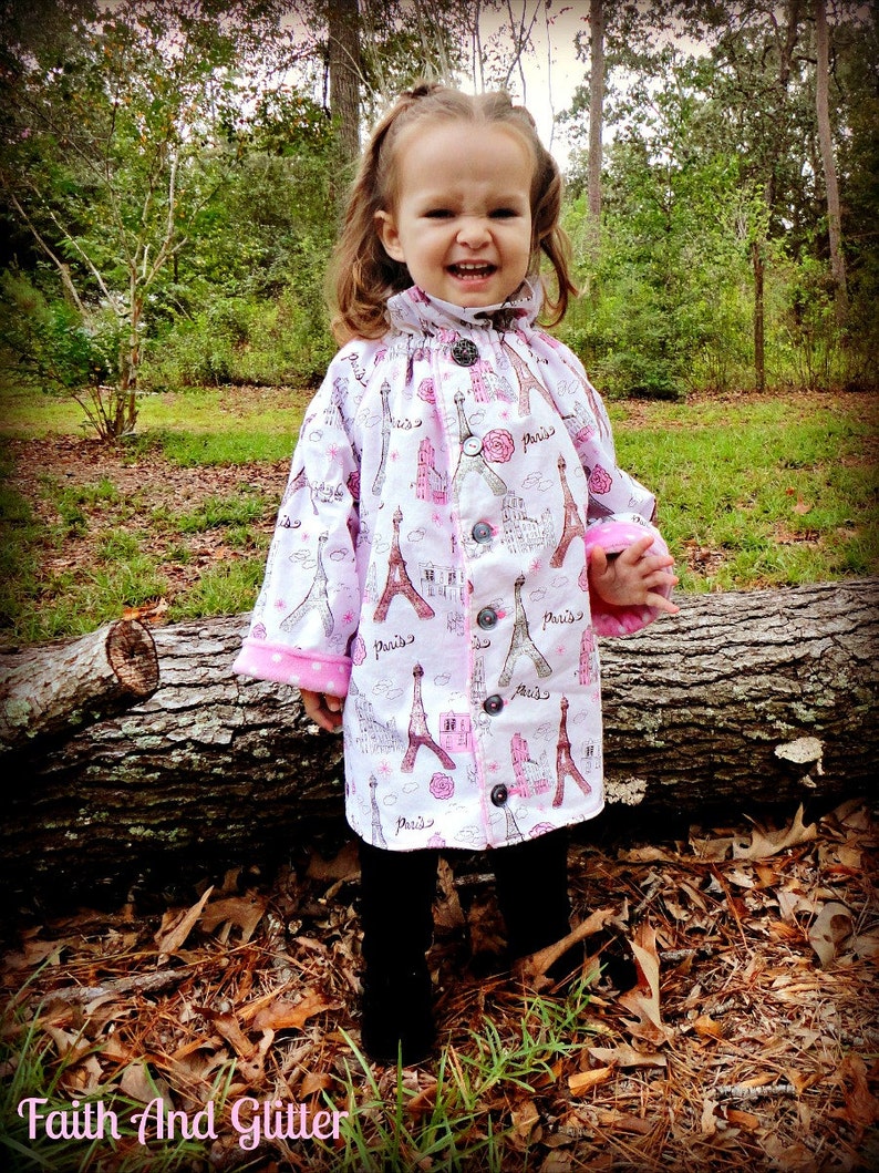 May include: A young girl wearing a white and pink jacket with a Paris theme. The jacket has a button closure and a pink trim. The girl is standing in front of a fallen log in a wooded area.