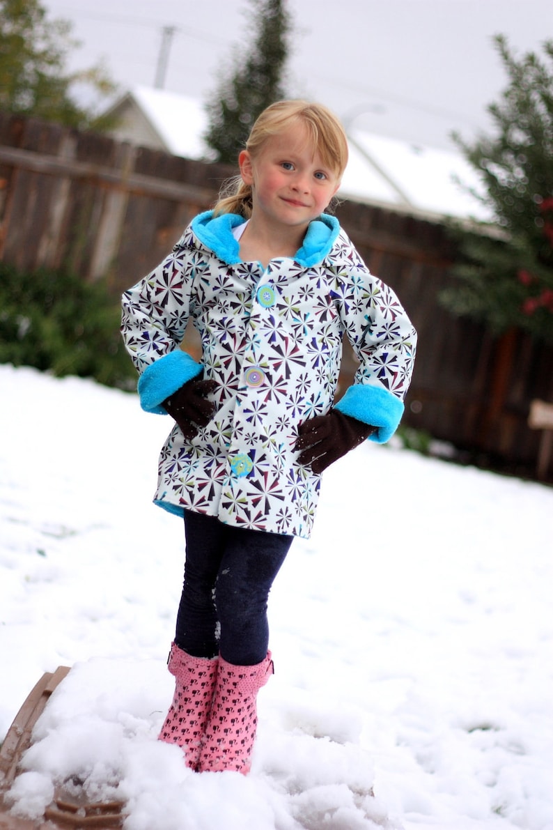 May include: A young girl wearing a white and black patterned jacket with blue trim and pink boots. She is standing in the snow.