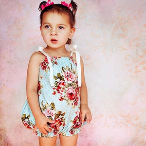 May include: A young girl wearing a blue and white floral print bubble romper with white straps. She has pink bows in her hair and is looking to the side.