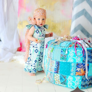 May include: A young child wearing a light blue and white key print romper with a pink and white headband stands in front of a colorful patchwork pouf. The pouf is turquoise, blue, and white with gold accents. The background is a pink and gold watercolor wash.