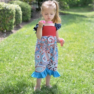 May include: A young girl wearing a red, blue, and white patterned dress with a ruffled hem blows bubbles with a pink wand. She is standing in a grassy area.