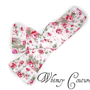 May include: White fabric with a floral pattern of red roses and green leaves. The fabric is folded and sewn into a scarf or headband shape. The text "Whimsy Couture" is written in cursive at the bottom right.