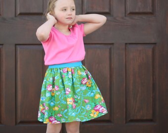 Puede incluir: Una niña pequeña con una camiseta rosa con una banda turquesa y una falda verde con flores. Lleva botas grises y está de pie frente a una puerta de madera.