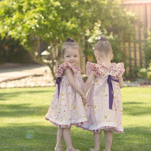 May include: Two young girls in matching pink dresses with ruffled sleeves and purple bows, holding hands in a sunny garden. The dresses have a polka dot pattern with colorful accents. They are wearing gold-colored shoes.