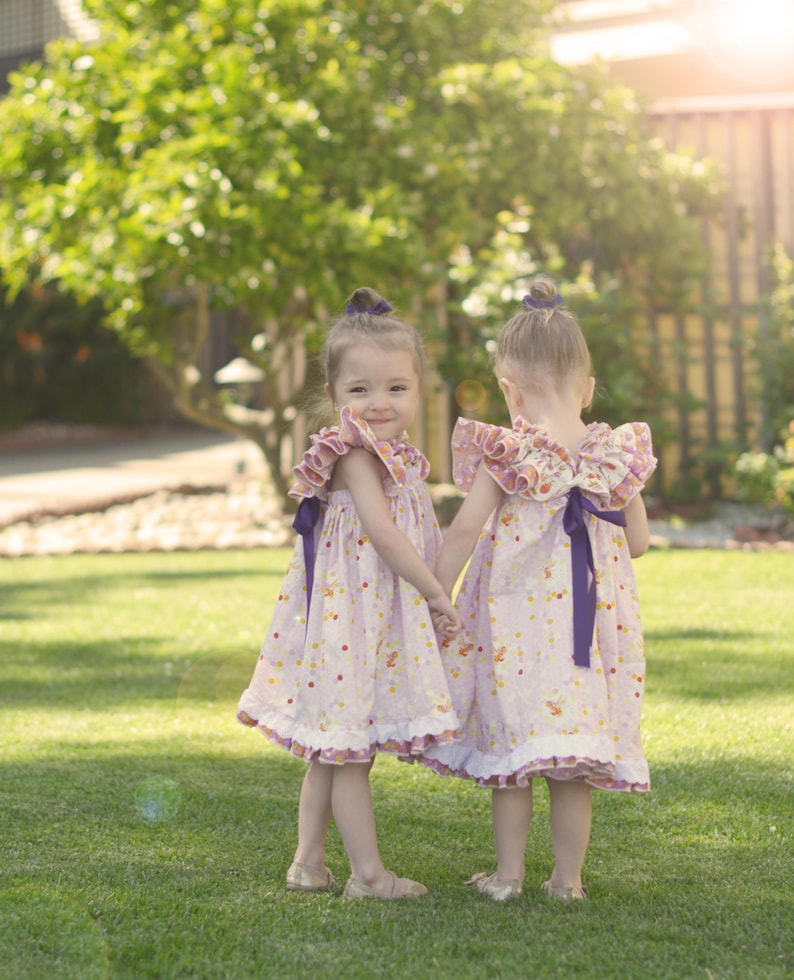 May include: Two young girls in matching pink dresses with ruffled sleeves and purple bows, holding hands in a sunny garden. The dresses have a polka dot pattern with colorful accents. They are wearing gold-colored shoes.