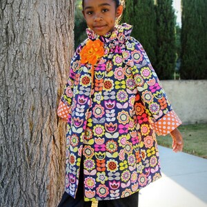 May include: A young girl wearing a colorful floral print jacket with orange flower accents and blue denim pants with ruffles at the bottom. The jacket has a ruffled collar and white polka dot trim on the sleeves.