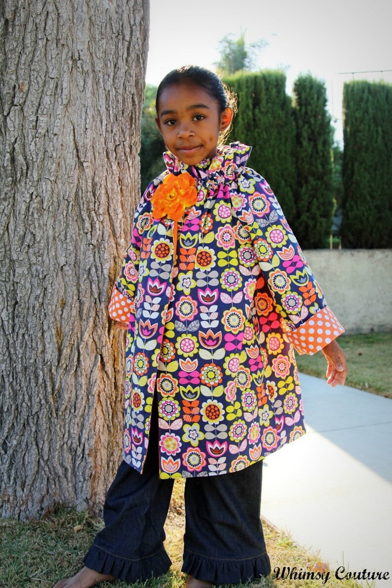 May include: A young girl wearing a colorful floral print jacket with orange flower accents and blue denim pants with ruffles at the bottom. The jacket has a ruffled collar and white polka dot trim on the sleeves.