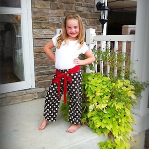 May include: A young person wearing a white shirt and black and white polka dot pants with a red bow tied at the waist. The person is standing on a concrete step in front of a brick wall and a white picket fence.