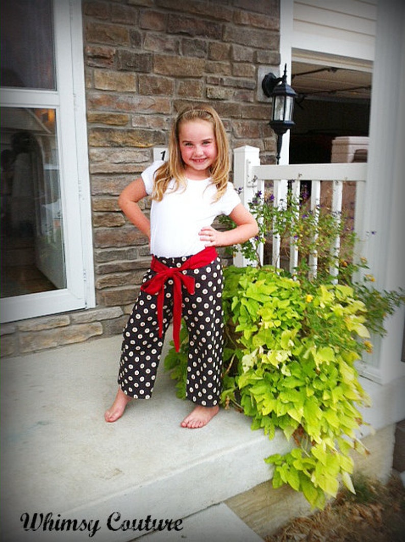 May include: A young person wearing a white shirt and black and white polka dot pants with a red bow tied at the waist. The person is standing on a concrete step in front of a brick wall and a white picket fence.