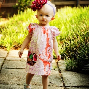 Puede incluir: Una niña pequeña con un vestido floral rosa y blanco y una diadema con una gran flor rosa. Está de pie en un camino de concreto con hierba verde en el fondo. El vestido tiene un bolsillo de parche con un diseño de búho.