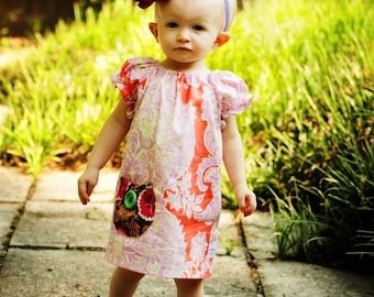 May include: A young girl wearing a pink and white floral dress with a large pink flower headband. She is standing on a concrete path with green grass in the background. The dress has a patch pocket with an owl design.