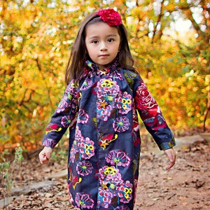 May include: A young girl wearing a dark blue floral print dress with a red flower headband. The dress has a ruffled collar and gold star accents. The girl is standing in a wooded area with fall foliage in the background.