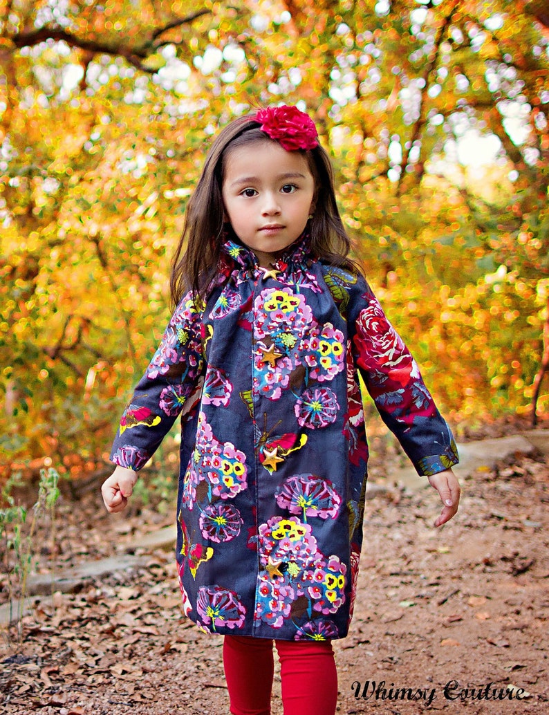 May include: A young girl wearing a dark blue floral print dress with a red flower headband. The dress has a ruffled collar and gold star accents. The girl is standing in a wooded area with fall foliage in the background.