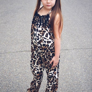 May include: A young girl wearing a black and brown leopard print jumpsuit with a black bow in her hair. She is standing on a gray asphalt surface and wearing black flip flops.