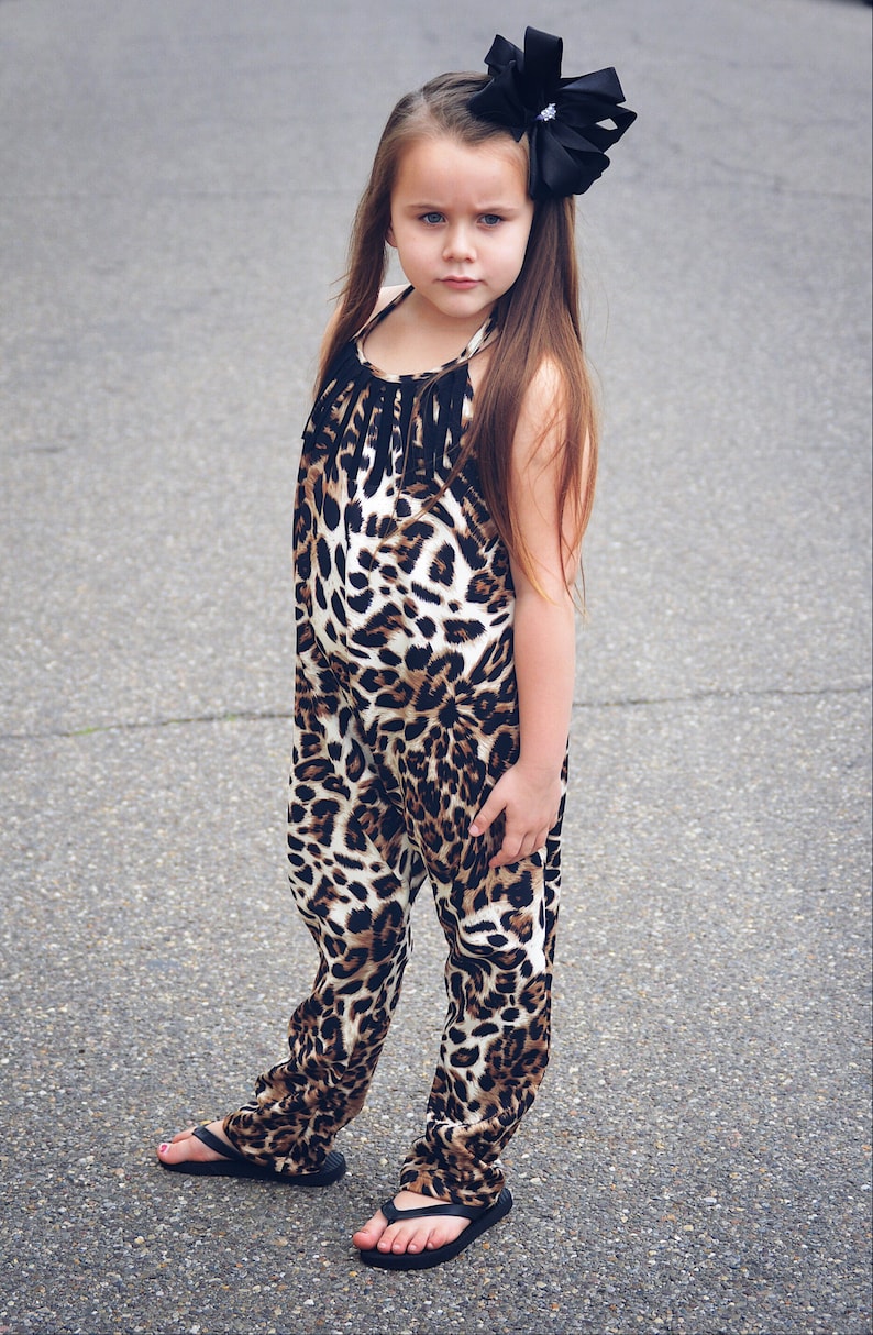 May include: A young girl wearing a black and brown leopard print jumpsuit with a black bow in her hair. She is standing on a gray asphalt surface and wearing black flip flops.