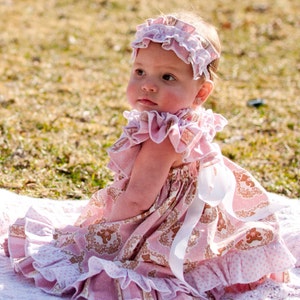 May include: A baby girl wearing a pink and white ruffled dress with a floral pattern and a matching headband. She is sitting on a white blanket in a grassy area.