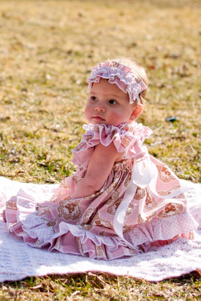 May include: A baby wearing a pink dress and matching headband, sitting on a white blanket outdoors. The dress features a repeating pattern with gold accents and ruffles. A white ribbon is tied at the back of the dress.
