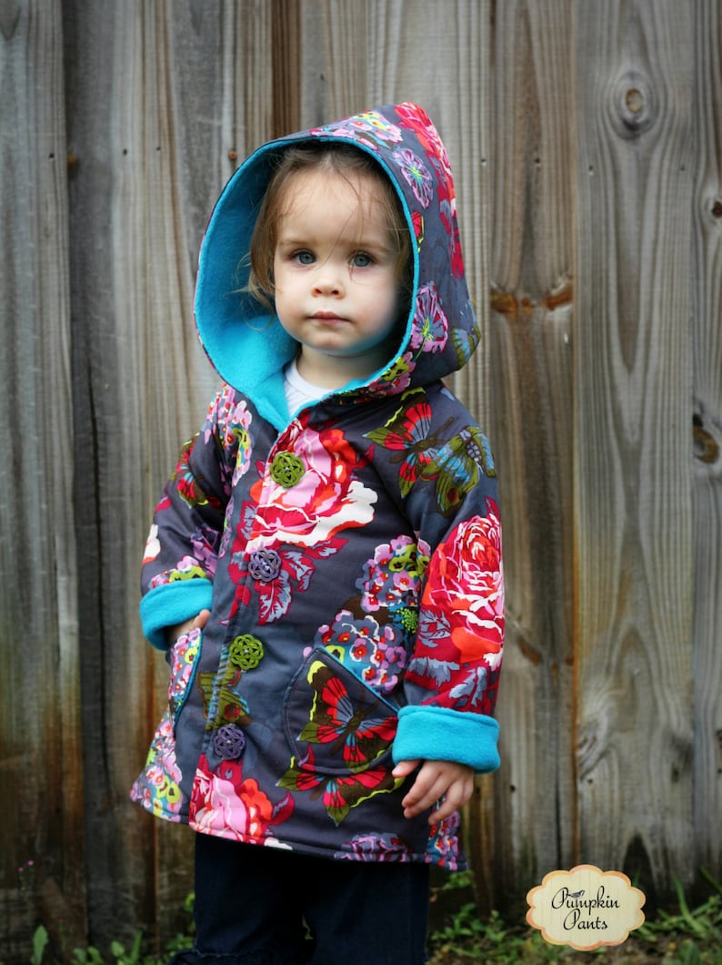 May include: A young child wearing a gray hooded jacket with a floral print and blue trim. The jacket has a large pocket on the front and buttons down the front. The child is standing in front of a wooden fence.