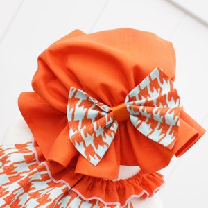 May include: An orange baby bonnet with a large bow featuring a light blue and orange fox pattern. The bonnet has a ruffled edge and is made of fabric. The bow is centered on the bonnet.