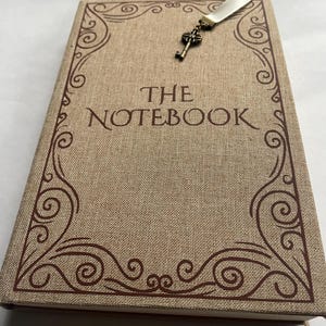 May include: A tan notebook with the words "THE NOTEBOOK" printed on the cover. The cover has a decorative brown border and a key charm attached to a white ribbon bookmark.