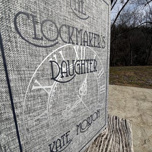 Peut inclure: Un livre à couverture rigide gris intitulé "The Clockmaker's Daughter" de Kate Morton. La couverture présente des lettres argentées et un motif de cadran d'horloge. Le livre repose sur une surface en bois vieilli, avec un arrière-plan extérieur flou.