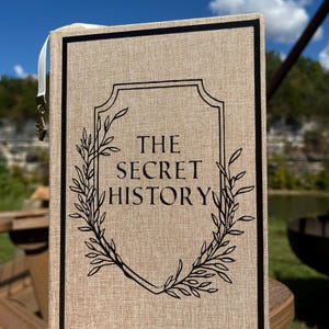 May include: A hardcover book titled "THE SECRET HISTORY" with a beige linen cover and black border. The title is in black, framed by a black decorative emblem and leafy branches. A white ribbon bookmark with a gold charm is attached.