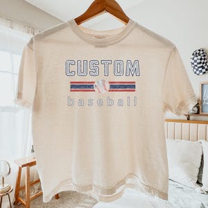 May include: Cream-colored t-shirt with the words "CUSTOM baseball" in a distressed font. The design includes a baseball graphic and red and blue stripes. The shirt is hanging on a wooden hanger.