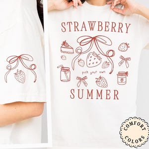 May include: Cream-colored t-shirt with a strawberry-themed design. The shirt features the words "STRAWBERRY SUMMER" and illustrations of strawberries, bows, a slice of cake, and a jar of jam. The left sleeve has a smaller strawberry and bow design.