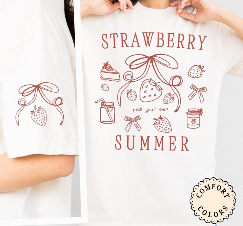 May include: Cream-colored t-shirt with a strawberry-themed design. The shirt features the words "STRAWBERRY SUMMER" and illustrations of strawberries, bows, a slice of cake, and a jar of jam. The left sleeve has a smaller strawberry and bow design.