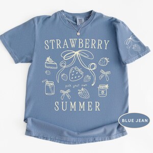 May include: A blue jean colored t-shirt with the words "STRAWBERRY SUMMER" in white script. The shirt features a strawberry and bow graphic design. The shirt has short sleeves and a crew neck.