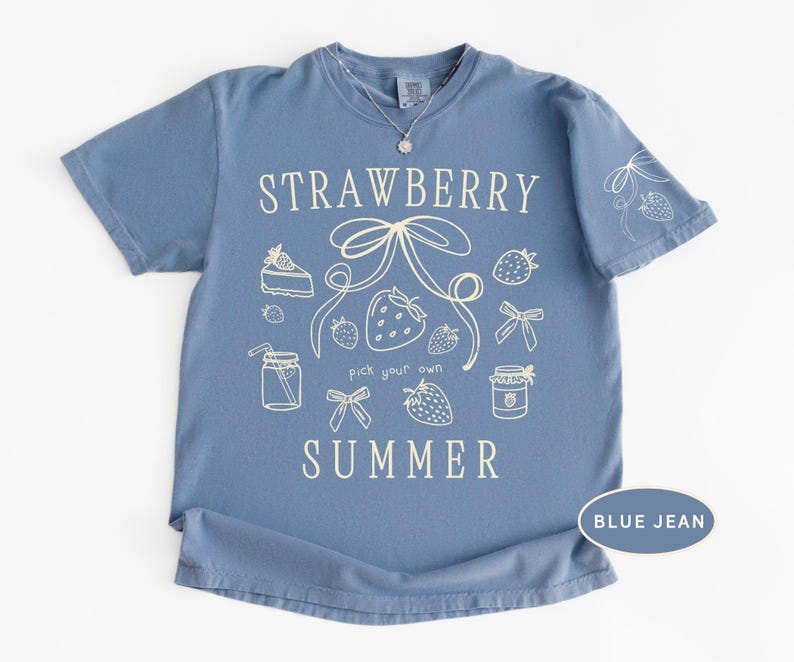May include: A blue jean colored t-shirt with the words "STRAWBERRY SUMMER" in white script. The shirt features a strawberry and bow graphic design. The shirt has short sleeves and a crew neck.