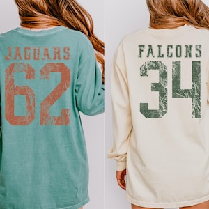 May include: Two long-sleeved shirts. One is teal with "JAGUARS" and the number "62" in distressed orange lettering. The other is cream-colored with "FALCONS" and the number "34" in distressed green lettering.