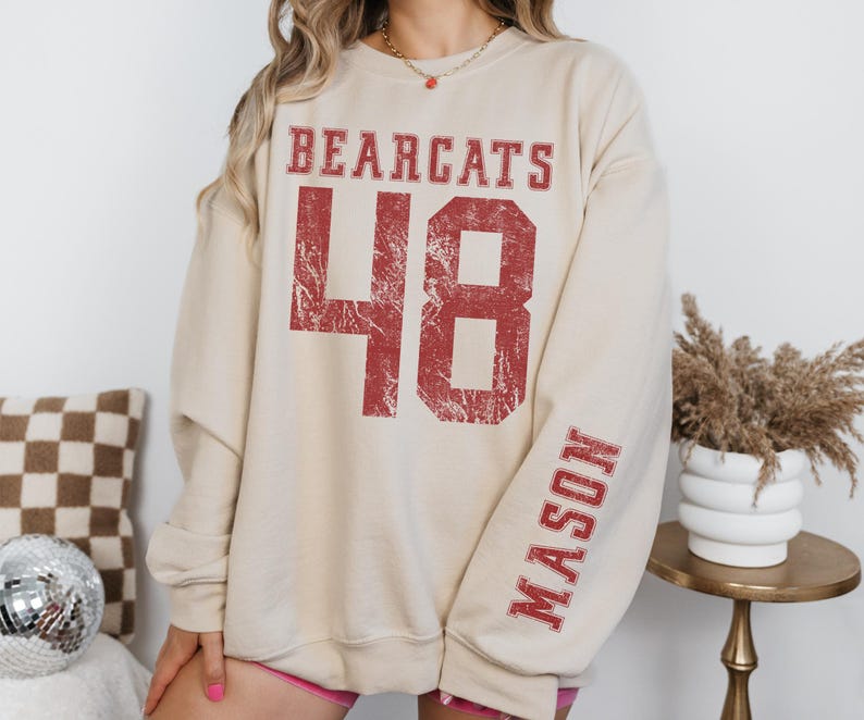 May include: Cream-colored sweatshirt with the word "BEARCATS" and the number "48" in distressed red lettering. The name "MASON" is printed vertically on the sleeve in the same red font. The sweatshirt has a relaxed fit.