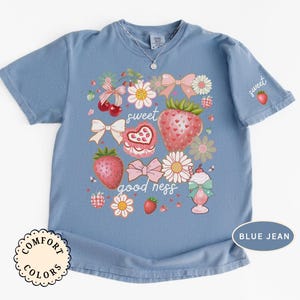 Op de afbeelding: Blauw Comfort Colors T-shirt met een aardbeienthema. Het ontwerp bevat aardbeien, strikken, bloemen en de woorden "sweet goodness". Een kleine aardbei staat op de mouw.