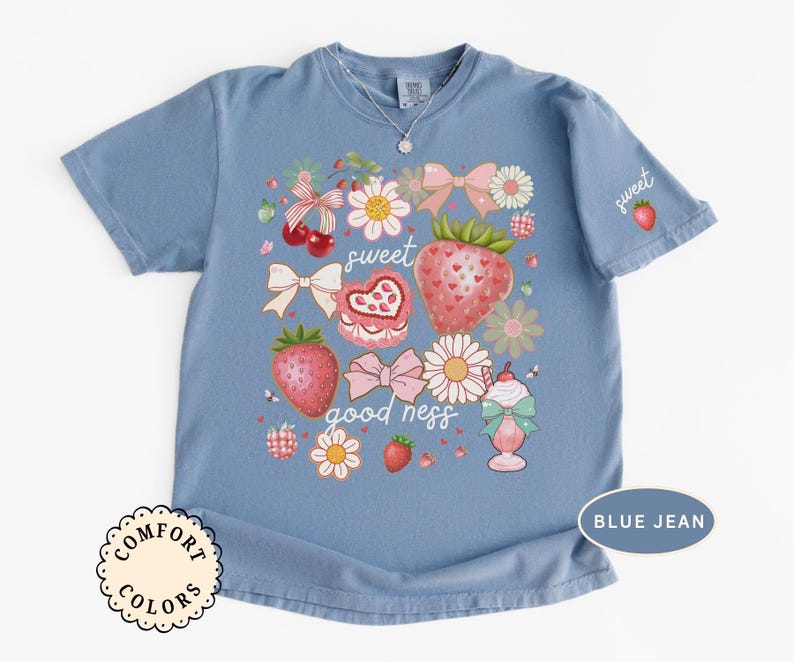 Op de afbeelding: Blauw Comfort Colors T-shirt met een aardbeienthema. Het ontwerp bevat aardbeien, strikken, bloemen en de woorden "sweet goodness". Een kleine aardbei staat op de mouw.