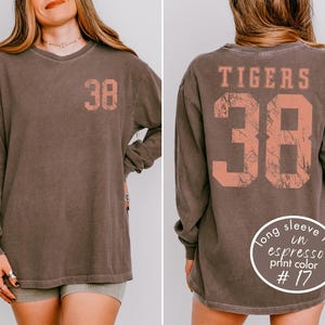 May include: A long-sleeve espresso-colored shirt with the number 38 printed on the front and back in a coral color. The word "TIGERS" is printed above the number on the back. The shirt has a vintage, worn look.