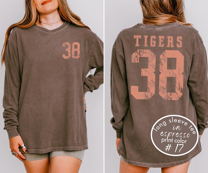 May include: A long-sleeve espresso-colored shirt with the number 38 printed on the front and back in a coral color. The word "TIGERS" is printed above the number on the back. The shirt has a vintage, worn look.