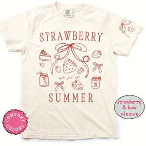 May include: Cream-colored t-shirt with a red strawberry and bow design. The shirt features the words "STRAWBERRY SUMMER" and the phrase "pick your own". The sleeve has a strawberry and bow design.