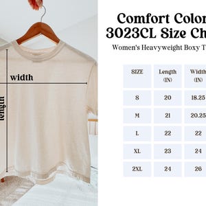 May include: A cream-colored, heavyweight boxy t-shirt hangs on a wooden hanger. The image includes a size chart with measurements in inches for sizes S to 2XL. The text on the chart reads "Comfort Colors 3023CL Size Chart" and "Women's Heavyweight Boxy T-Shirt."