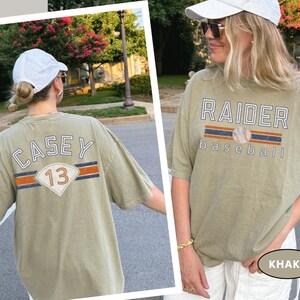 May include: Khaki-colored t-shirts with baseball-themed graphics. One shirt features "RAIDER baseball" with a baseball graphic. The other shirt displays "CASEY 13" with a baseball-style design. Both shirts have short sleeves and a relaxed fit.