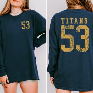 May include: Navy blue long-sleeve shirt with the number "53" in distressed gold on the front and back. The back of the shirt also features the word "TITANS" above the number. The shirt is a comfortable, casual style.
