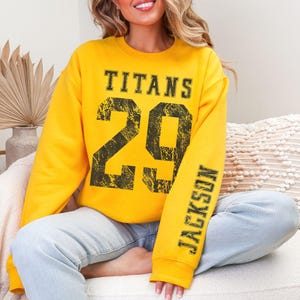 May include: A yellow sweatshirt with the word "TITANS" above the number "29" in black distressed lettering. The sleeve reads "JACKSON" in black. The sweatshirt is worn with light blue jeans.