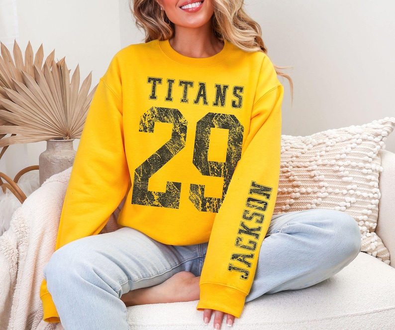 May include: A yellow sweatshirt with the word "TITANS" above the number "29" in black distressed lettering. The sleeve reads "JACKSON" in black. The sweatshirt is worn with light blue jeans.
